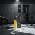 Maison Alhambra Dark Door Intense by Maison Alhambra 100ml Men — A clear glass perfume bottle with yellow liquid sits on a wet concrete floor in a locker room setting, with lockers and gym equipment in the background.