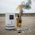 Maison Alhambra Ebene by Maison Alhambra Perfume 90 ml EDP — Cracked glass perfume bottle with colorful smoke rising, next to its white box on a concrete surface with broken glass shards.