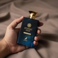 Maison Alhambra Ethnic Oud by Maison Alhambra 100ml EDP — A hand holds a dark blue glass perfume bottle with a gold cap and gold lettering, set against a soft brown fabric background.