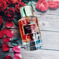 Maison Alhambra Exclusif Rose Maison Alhambra 100ml EDP — A clear glass perfume bottle with a gold cap, filled with amber-colored liquid, rests on a wooden surface surrounded by red rose petals and leaves.
