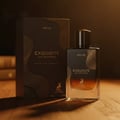 Maison Alhambra Exquisite Prive Maison Alhambra 100 ml EDP Men — A dark brown perfume box and a clear glass bottle with a dark cap, both featuring gold wave patterns and the brand's deer logo, set on a wooden surface with soft lighting.