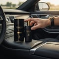 Maison Alhambra Glacier Le Noir by Maison Alhambra 100ml Men — A hand holds a black perfume bottle with a gold cap, next to its matching cylindrical packaging, inside a car.