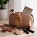 Maison Alhambra Glossy By Maison Alhambra 100ml Women EDP — A quilted pink makeup bag with makeup brushes, a bottle of Glossy perfume, eyeshadow palettes, lipstick, and a compact powder on a wooden surface.