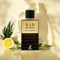 Maison Alhambra B.A.D. Homme Eau de Parfum — A dark glass perfume bottle with a gold label, black textured cap, and a deer logo, placed with a lemon slice and green sprigs on a reflective surface.