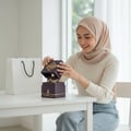Maison Alhambra Infini Rose by Maison Alhambra 100ml EDP Unisex — Wanita bertudung membuka kotak hadiah heksagon beraksen emas, menampakkan botol minyak wangi di dalamnya, beg kertas putih di sisinya.