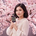 Maison Alhambra Infini Rose by Maison Alhambra 100ml EDP Unisex — Wanita muda pegang botol minyak wangi ungu corak geometri emas di depan pokok bunga sakura merah jambu.