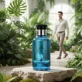 Maison Alhambra Jean Lowe Azure by Maison Alhambra EDP 100 ml — Blue glass perfume bottle with black cap and silver accents, placed on a textured stone with moss, in a lush green botanical garden setting with a man walking in the background.