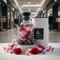 Maison Alhambra Jean Lowe Matiere by Maison Alhambra 100ml — A shattered glass perfume bottle with red and pink roses inside, surrounded by pink cyclamen flowers and a white cylindrical box in a mall setting.