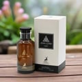 Maison Alhambra Jean Lowe Noir — Amber glass perfume bottle with black cap and gold accents, next to its white and black box with a deer logo, on a wooden surface with blurred pink flowers in the background.