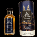 Maison Alhambra Jean Lowe Nouveau — A clear glass perfume bottle with golden liquid and a dark blue label featuring silver leaf motifs and a deer silhouette, placed next to a cylindrical perfume box with a night cityscape and twin towers illustration.