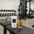 Maison Alhambra Jean Lowe Nouveau — A perfume bottle and its cylindrical packaging sit on a black gym bench, with dumbbells and gym equipment in the background.