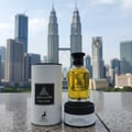 Maison Alhambra Jean Lowe Nouveau — A bottle of yellow perfume with a black cap sits on a black base, next to its white cylindrical box, with the Petronas Twin Towers and city skyline in the background.