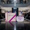 Maison Alhambra Just Married by Maison Alhambra Woman 100ml — A perfume bottle and its box displayed on a reflective surface, with a blurred mall background.