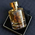 Maison Alhambra Karat by Maison Alhambra 100ml EDP — A clear glass perfume bottle with gold intricate design and a gold cap, resting on a black and gold box, with a black tassel.
