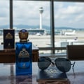 Maison Alhambra Kingsman by Maison Alhambra – 100 ml for Him, a blue perfume bottle with a gold crown cap, placed on a passport with sunglasses, with an airport runway view in the background.