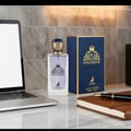Maison Alhambra Kingsman by Maison Alhambra – 100 ml for Him, a clear glass perfume bottle with a gold crown-like cap, sits next to its navy blue box on a marble desk with a laptop and a leather-bound notebook.