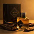 Maison Alhambra Kismet Magic by Maison Alhambra 100ml EDP Unisex — Black textured box with gold geometric pattern and 'Kismet Magic' text, alongside a clear glass perfume bottle with gold ribbed cap and a black label. Two pieces of dark, textured wood are placed in the foreground.