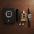 Maison Alhambra Kismet For Men Eau de Parfum — Black gift box with gold glitter, perfume bottle with gold cap, leather case, and brown leather watch on a wooden surface.
