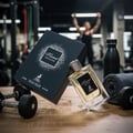 Maison Alhambra Kismet Moscow by Maison Alhambra 100ml EDP — A glass perfume bottle and its black box sit on a wooden surface with dumbbells and a rolled towel in a gym setting, with a blurred person lifting weights in the background.