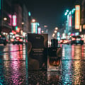 Maison Alhambra Exquisite Prive Maison Alhambra 100 ml EDP Men — A perfume bottle and its box sit on a wet, reflective street at night, with blurred city lights and traffic in the background.