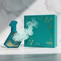 Maison Alhambra Luxe Emerald Perfume Maison Alhambra 100ml Unisex — Teal spray bottle with gold geometric pattern, releasing a cloud of mist, next to its matching teal gift box with gold accents, on a marble surface.