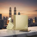 Maison Alhambra Luxe Gold — Botol dan kotak minyak wangi emas dipamerkan di atas permukaan marmar dengan latar belakang langit bandar dan pencakar langit semasa matahari terbenam, pasport juga kelihatan.