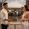 Maison Alhambra Luxe Gold – Lelaki berbaju krim dan wanita berbaju picang tersenyum antara satu sama lain di atas meja marmar memaparkan kotak dan botol wangian emas, dengan lampu bokeh di latar belakang.