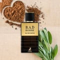 Maison Alhambra B.A.D. Homme Eau de Parfum — A dark brown glass perfume bottle with a gold label and black cap, surrounded by cocoa powder in a heart-shaped dish and fresh sage leaves.