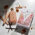 Maison Alhambra Decadent Dream — Peach-toned perfume bottle with gold cap, surrounded by wood sticks, vanilla pods, amber resin pieces, and shattered glass on a marble surface.