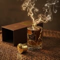 Maison Alhambra Exclusif Tabac by Maison Alhambra – 100 ml Unisex, a glass perfume bottle with golden cap and smoke rising, next to its brown box on a woven mat.