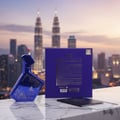 Maison Alhambra Luxe Nightlife Unisex 100 ml — Blue metallic perfume bottle with white starry design and gold cap, placed on a marble surface with a passport and boarding pass in front of a city skyline with Petronas Towers at dusk.