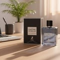 Maison Alhambra Maitre De Blue — Botol minyak wangi kaca jernih bertutup hitam dan tulisan emas, bersebelahan kotak hitam emasnya, di atas meja kayu bersama komputer riba dan pokok dalam pasu.