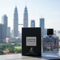 Maison Alhambra Maitre De Blue Eau de Parfum — Botol minyak wangi kaca jernih bertutup hitam dan kotak hitam bertulis putih, berlatar belakang pemandangan bandar yang kabur dengan pencakar langit tinggi.