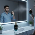 Maison Alhambra Glacier Bold by Maison Alhambra 100ml EDP — Man spraying perfume in front of a mirror with a backlit frame, reflection shows the spray mist.