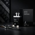 Maison Alhambra Man Black Edition — Black perfume bottle and box with silver cap, alongside a watch and cufflinks on a dark textured surface, with a patterned fabric in the background.