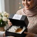 Maison Alhambra Montaigne Coco Maison Alhambra 100ml EDP — Wanita bertudung dan berbaju kilat membuka kotak hadiah hitam berisi botol minyak wangi kaca di dalam kertas carik putih.