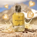 Maison Alhambra Montaigne Vanille 100ml — A clear glass perfume bottle with yellow liquid, surrounded by latte art hearts, milk splashes, vanilla pods, and coffee beans, with a mountain landscape background.