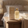 Maison Alhambra Musk Vanille EDP — A bottle of Musk Vanille by Maison Alhambra perfume and its box sit on a wooden nightstand next to a knitted blanket, with a bed in the background.