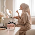 Maison Alhambra My Party Maison Alhambra 100ml EDP Woman — A clear glass perfume bottle with pink liquid and a black ribbon bow sits on a wooden table. A woman in a hijab applies makeup in the blurred background.