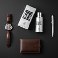 Maison Alhambra No.2 Men by Maison Alhambra 80ml EDP — Silver perfume bottle and box with black text, a brown leather watch, a brown leather wallet, and a silver pen on a dark background.