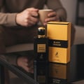 Maison Alhambra Black Origami — Botol minyak wangi hitam dengan penutup emas di sebelah kotak kuning dan hitamnya, diletakkan di atas permukaan hitam berkilat. Seseorang memegang cawan kelihatan kabur di latar belakang.