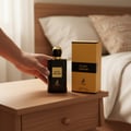 Maison Alhambra Black Origami — Tangan memegang botol minyak wangi hitam berpenutup emas, diletakkan di atas meja sisi katil kayu bersebelahan kotak emas dan hitamnya. Katil kelihatan di latar belakang.