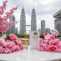 Maison Alhambra Paris Garden Maison Alhambra 100ml EDP Woman — A bottle of Maison Alhambra Paris Garden perfume sits on a marble surface, surrounded by pink cherry blossoms and roses, with the Petronas Twin Towers in the background.