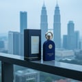 Maison Alhambra Philos Pura by Maison Alhambra 100ml EDP Man — A navy blue perfume bottle with a gold cap and a matching navy blue box are displayed on a balcony railing, with the Petronas Twin Towers in the blurred background.
