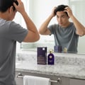 Maison Alhambra Philos Pura by Maison Alhambra 100ml EDP Man — A young man styles his hair in front of a mirror, with a purple perfume bottle and its box displayed on a granite countertop.