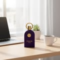 Maison Alhambra Philos Pura by Maison Alhambra 100ml EDP Man — A deep purple perfume bottle with a gold ring and emblem sits on a wooden desk next to a laptop and a white coffee mug.