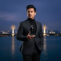 Maison Alhambra Glacier Bold by Maison Alhambra 100ml EDP — Man in a suit holding a dark blue perfume bottle with a silver cap, standing by a river with a bridge and city lights in the background at dusk.