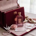 Maison Alhambra Rose Origami — A maroon velvet handbag is open, revealing its interior. A clear glass perfume bottle with a gold cap sits in front of it, next to a pink origami rose, a white origami crane, and a rose quartz bracelet.
