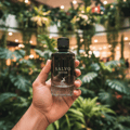 Maison Alhambra Salvo Intense by Maison Alhambra 100ml EDP — A hand holds a dark glass perfume bottle with a black cap, featuring the 'SALVO INTENSE' label and a deer logo, against a blurred background of lush green plants and warm lights.
