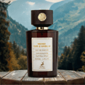 Maison Alhambra Smoked Oud & Vanilla — Amber glass perfume bottle with wooden cap and gold emblem, set on a rustic wooden table with a blurred mountain forest background.